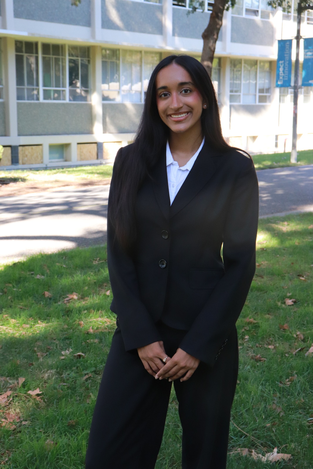 Iqra Ahmad stands on campus wearing a black suit with a white oxford.