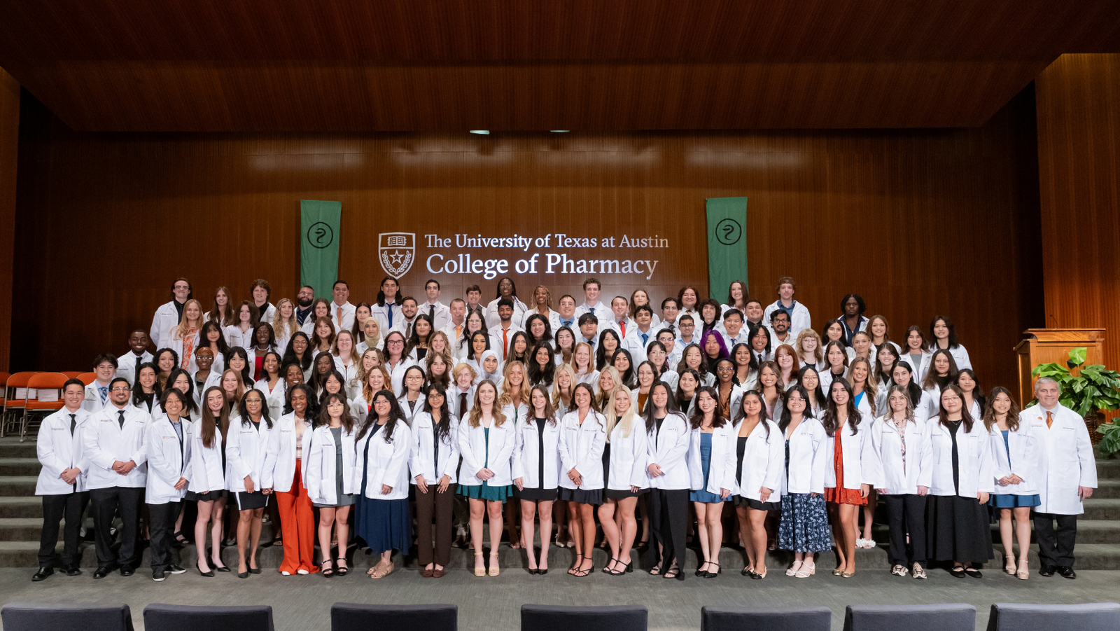 A large group of pharmacy students wearing white coats stands on a stage in front of a backdrop that reads "The University of Texas at Austin College of Pharmacy.