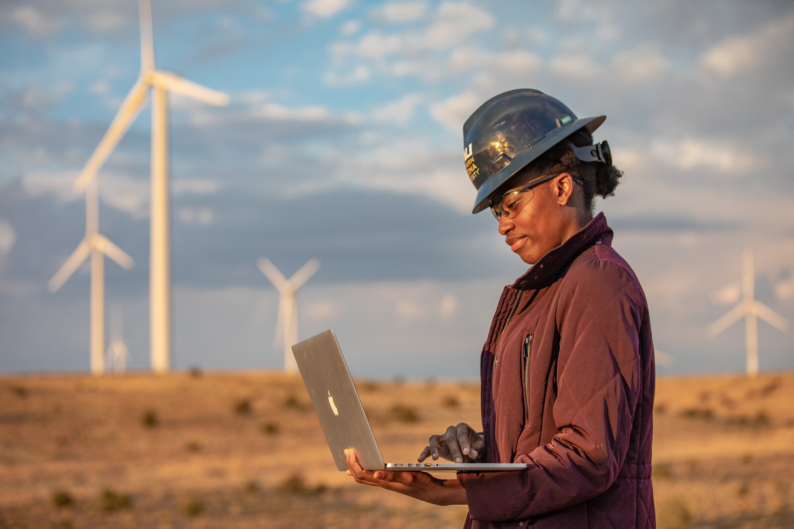 An engineering student checks her computer. In the background, there are numerous windmills.