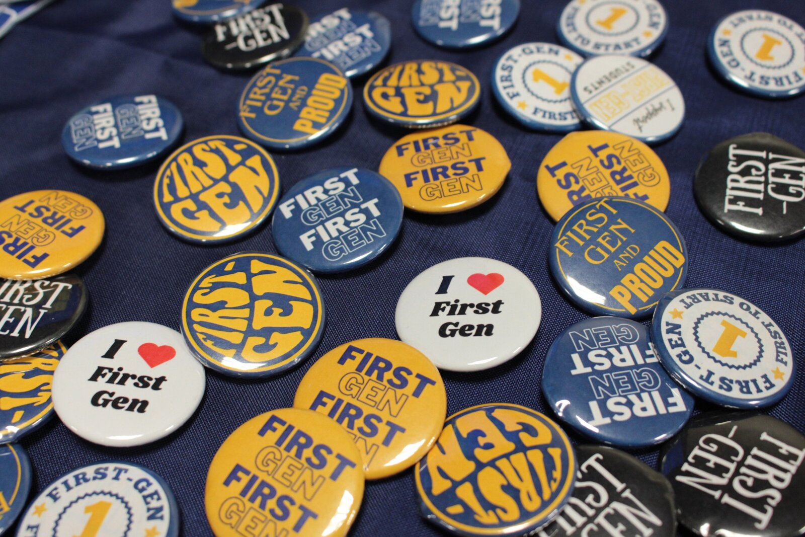 A collection of different pins are laid out on a table. They read: "First Gen and Proud", "I Heart First Gen".