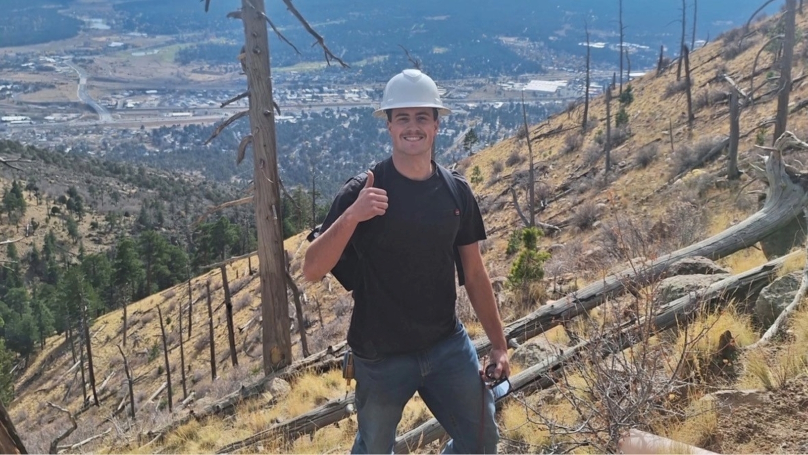 NAU student Ralph Testa, the first Dean's Distinguished Fellow from Summer 2026, gives a thumbs up. He is on top of a mountain with a hard hat on.