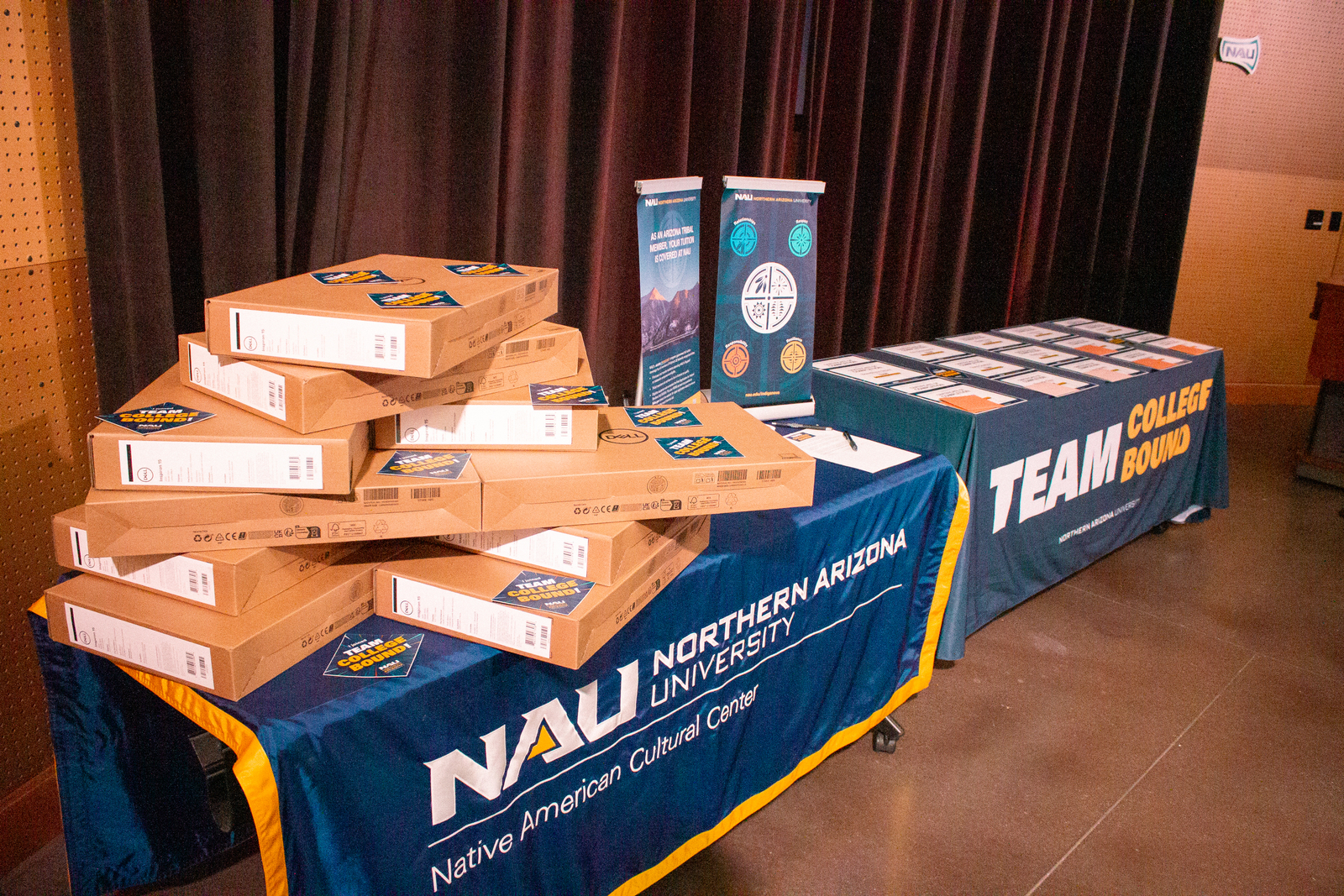 A table with a banner that reads, "NAU Native American Cultural Center" has a pile of Dell laptops on it. A table with a banner that reads, "NAU Native American Cultural Center" has a pile of Dell laptops on it.